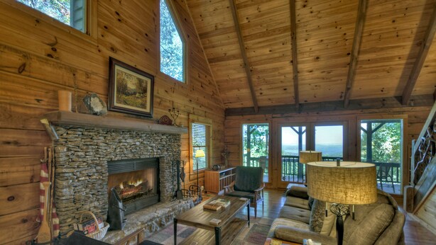 Interior acogedor de una cabaña con chimenea de piedra, paredes de madera, un espacio de estar confortable y ventanas amplias con vistas al exterior.