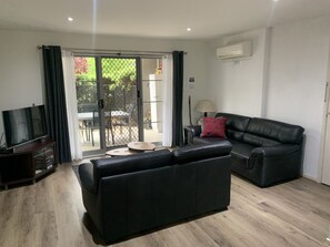 A modern living room with a black leather sofa, a TV on a wooden stand, and a sliding glass door leading to an outdoor patio with a table and chairs.