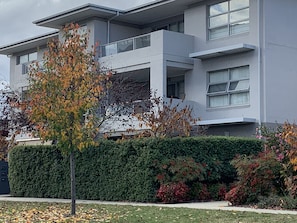 A modern two-story building with a balcony, surrounded by well-maintained hedges and trees with autumn leaves.