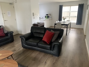 A modern living room with a black leather sofa, a wooden coffee table, and a dining area with a white table and chairs.