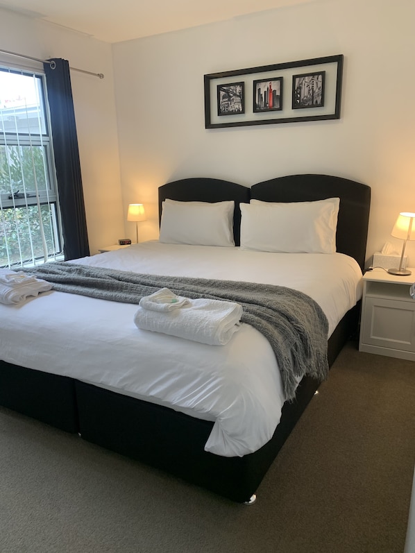 A neatly made bed with white linens, a grey blanket, and folded towels. There are two bedside lamps, a framed picture above the bed, and a window with curtains.