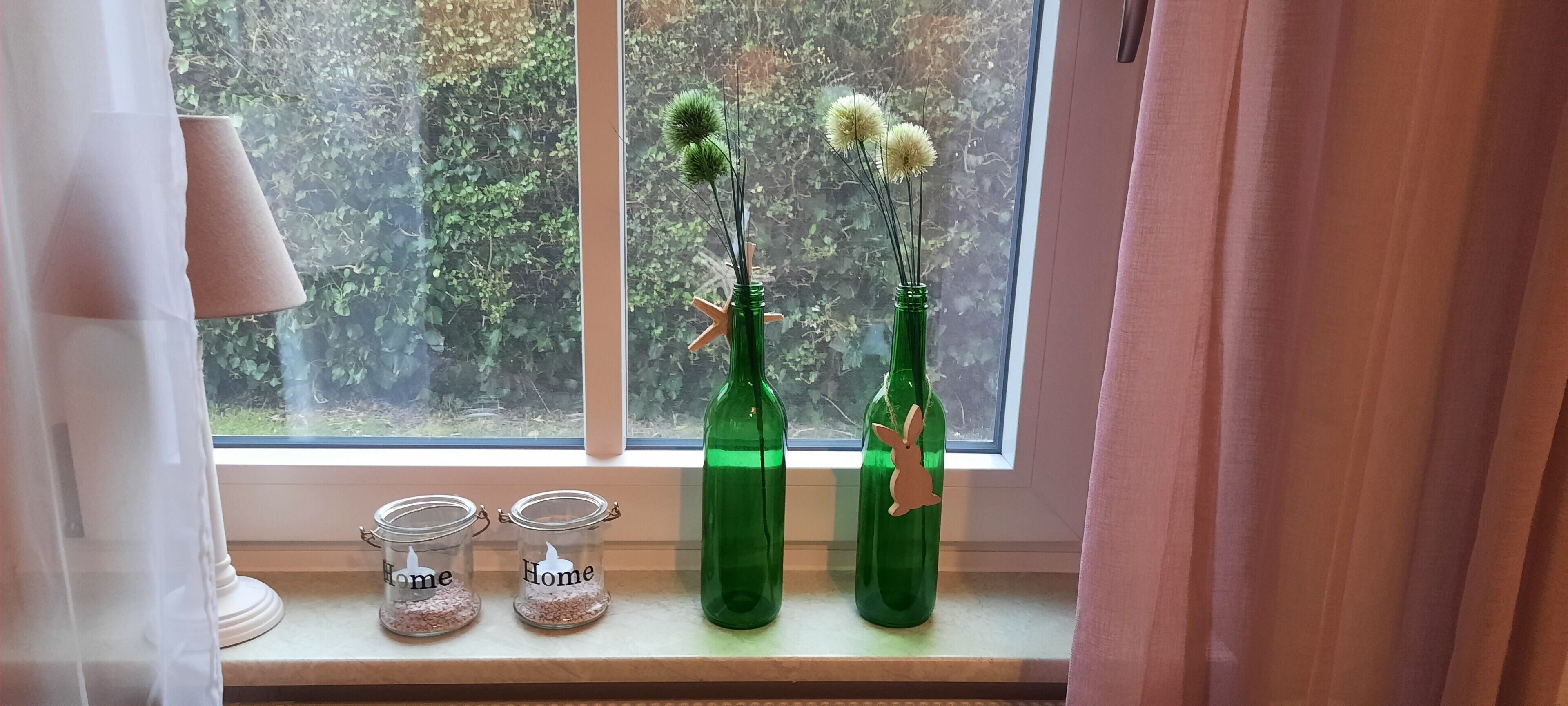 Uma janela com duas garrafas de vidro verde, cada uma decorada com um coelho, e dois potes de vidro com a palavra 'Home'.