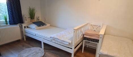 A bedroom with a single bed, a smaller bed, a radiator, a window with curtains, a plant, and a small rug.