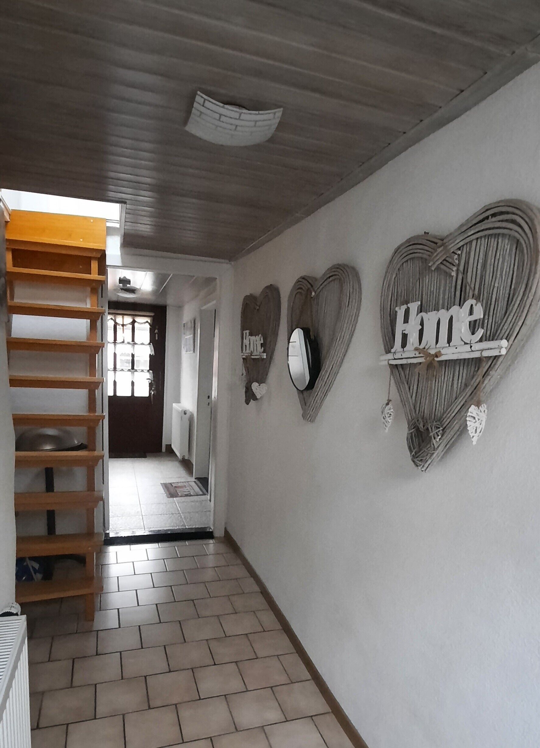 Ein Flur mit einer Holzleiter, einem Kachelboden und Wanddekorationen in Herzform, auf einer davon ist das Wort 'Home' zu sehen.
