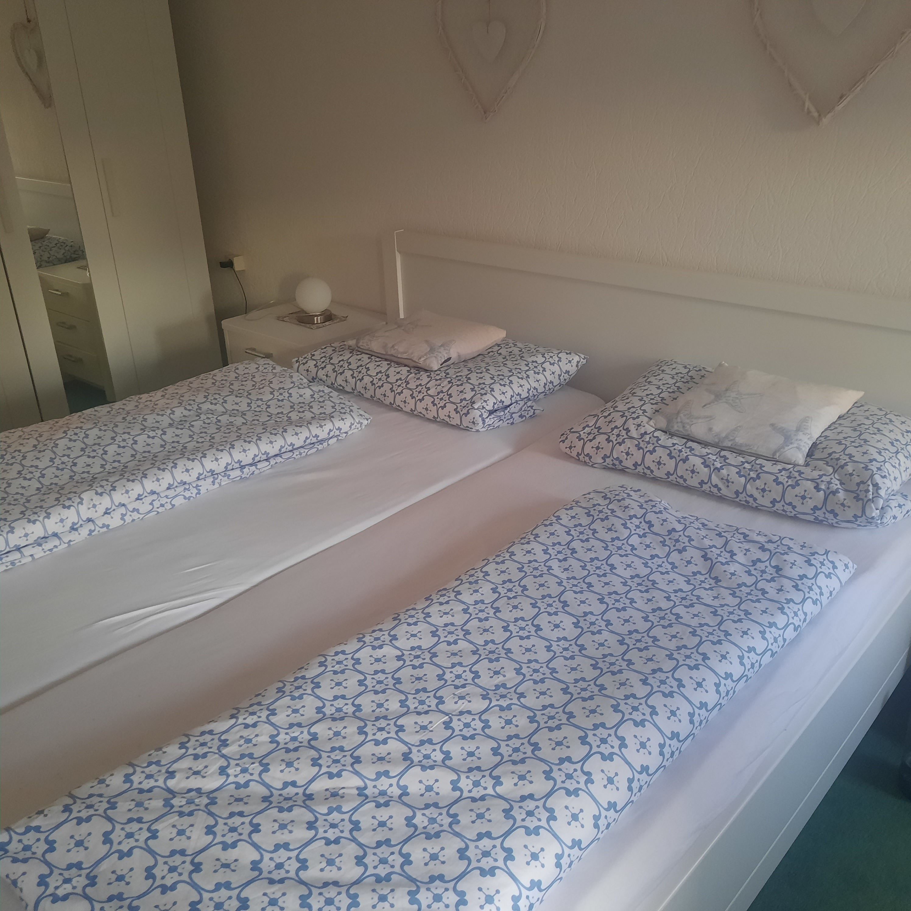 Una camera da letto con due letti singoli, ognuno con biancheria e cuscini a motivi decorativi, un comodino con una lampada e una decorazione a forma di cuore appesa al muro.