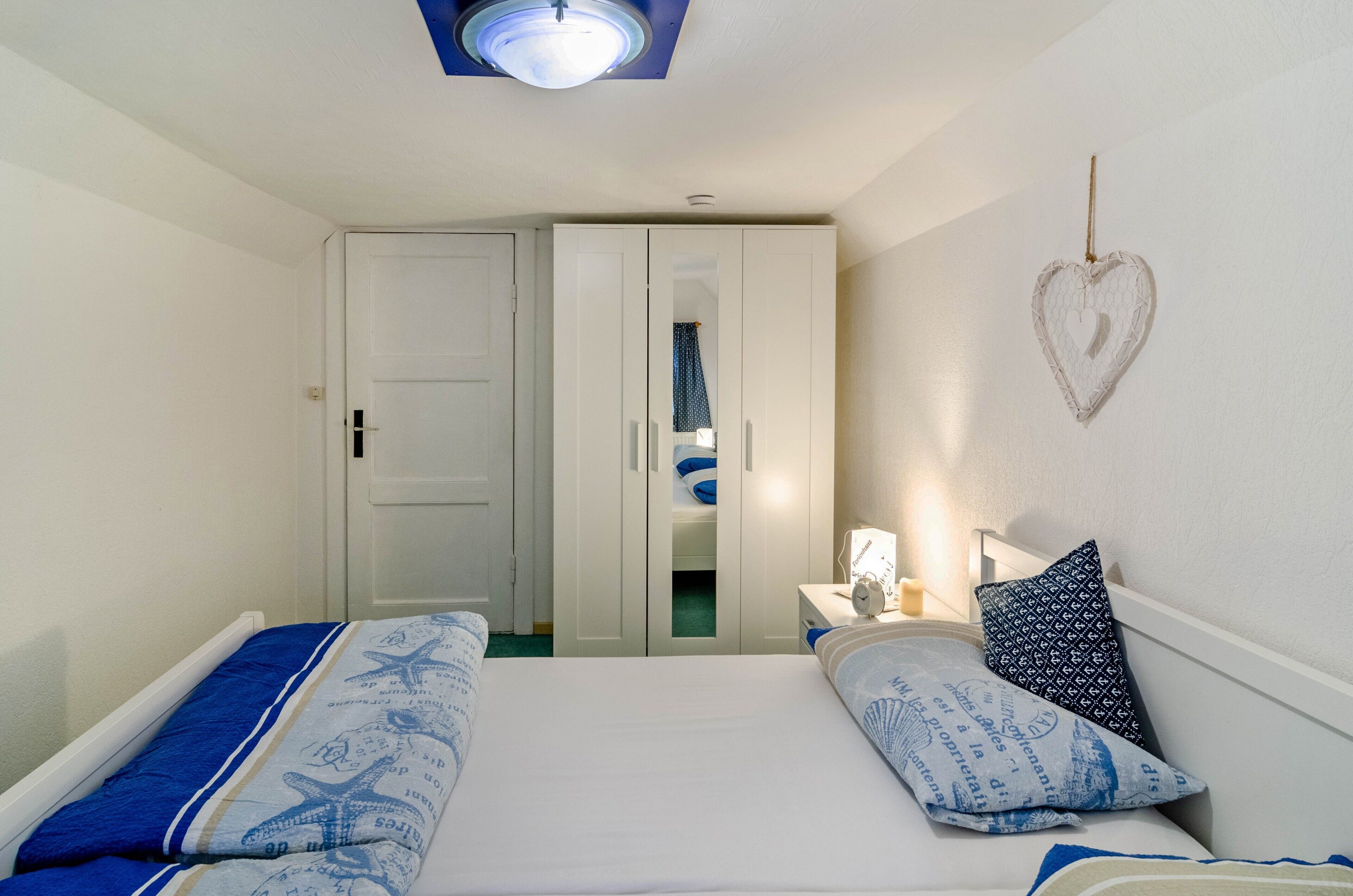 A bedroom with two beds, a wardrobe, a bedside table with a lamp, and a heart-shaped decoration on the wall.