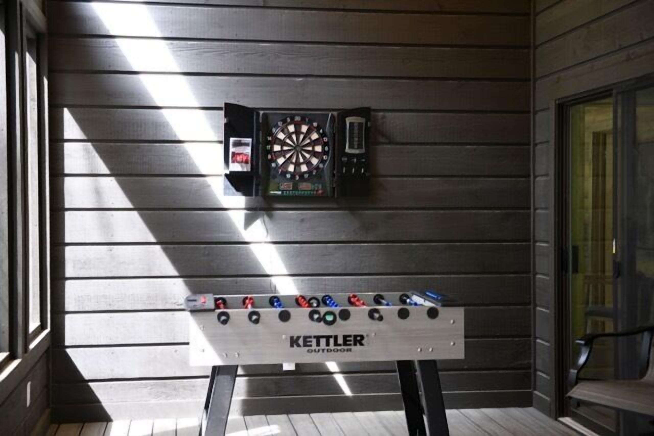 Una taula de futbolí amb el lema "KETTLER OUTDOOR" i un panell de dardo penjat a la paret.