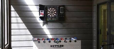 Una taula de futbolí amb el lema "KETTLER OUTDOOR" i un panell de dardo penjat a la paret.