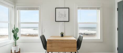 Une pièce avec une table en bois et deux chaises, de grandes fenêtres donnant sur l’océan et un tableau encadré au mur.