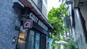 Une rue étroite bordée d’un immeuble portant l’inscription « COZY RIVERVIEW », avec une façade moderne, de grandes fenêtres et un petit espace extérieur aménagé avec des parasols.