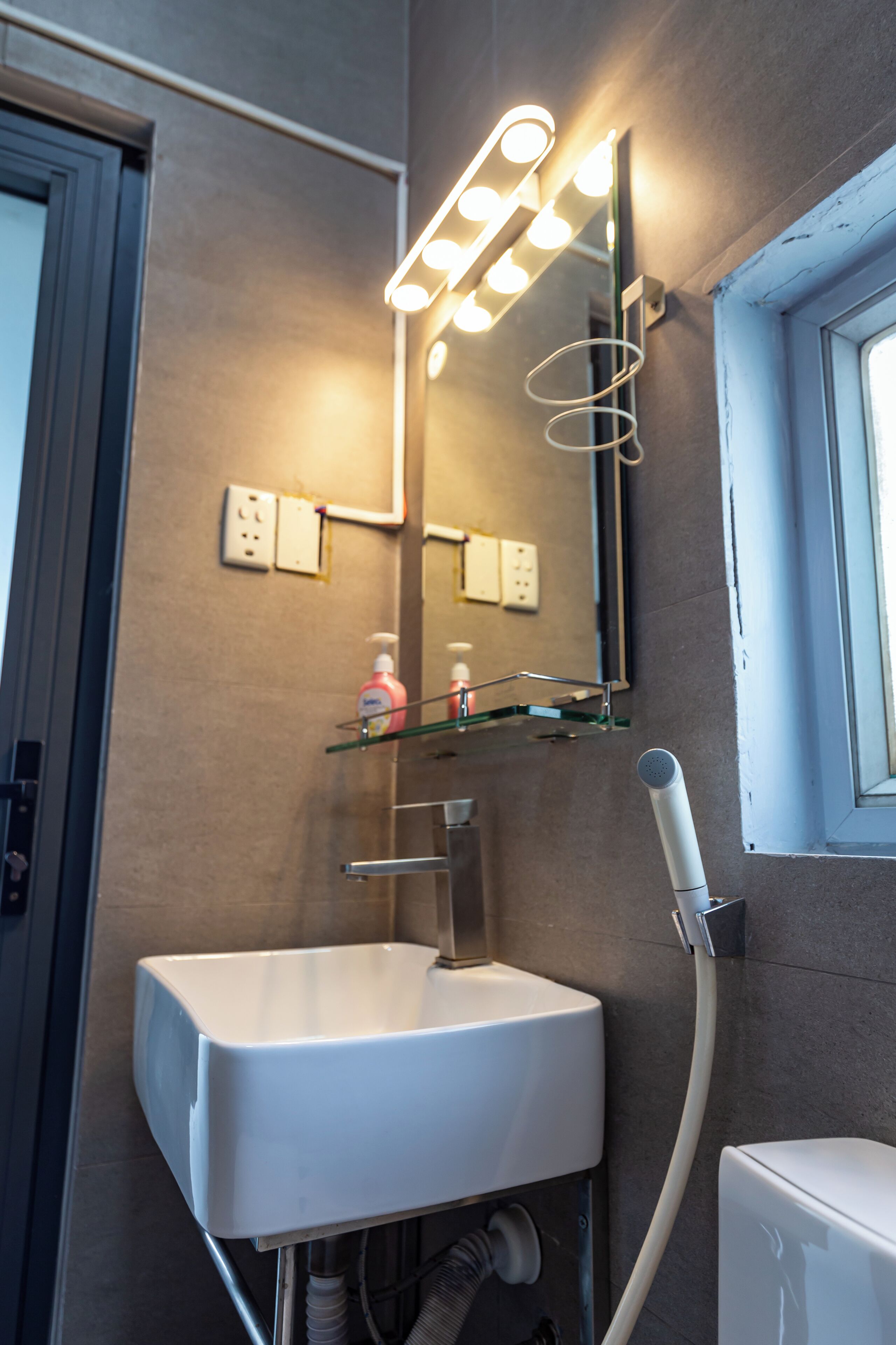 Un bagno con un lavandino bianco, uno specchio illuminato, un soffione doccia a mano e una finestra.