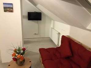A room with a red sofa, a wooden side table with a flower vase, a flat-screen TV mounted on the wall, and a radiator.