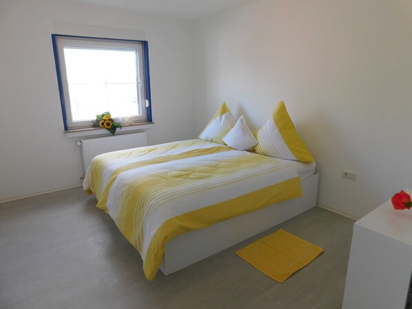 A bedroom with a bed, a window, a radiator, and a small table with a red object on it.