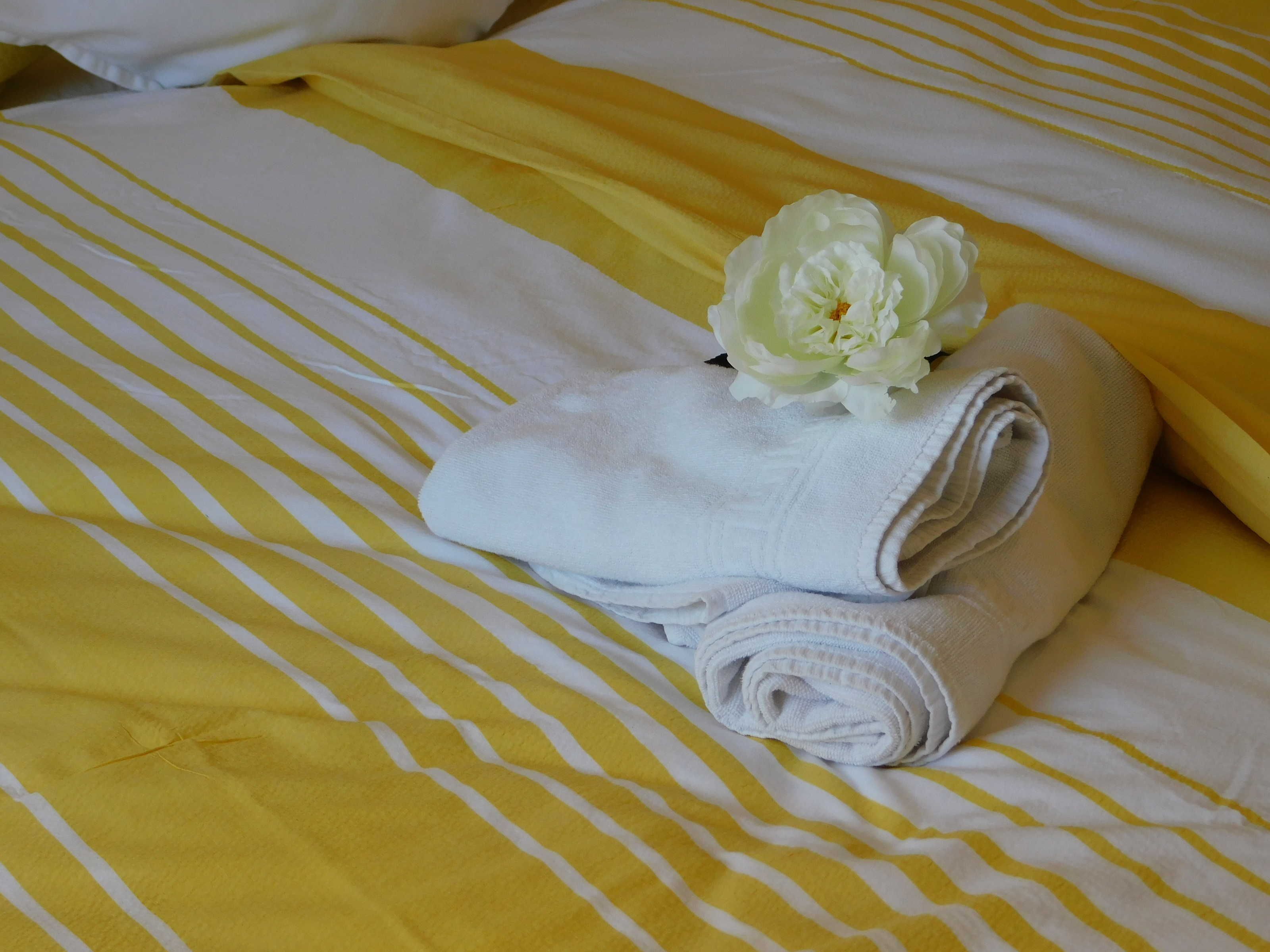 A bed with yellow and white striped bedding, a rolled white towel, and a white flower.