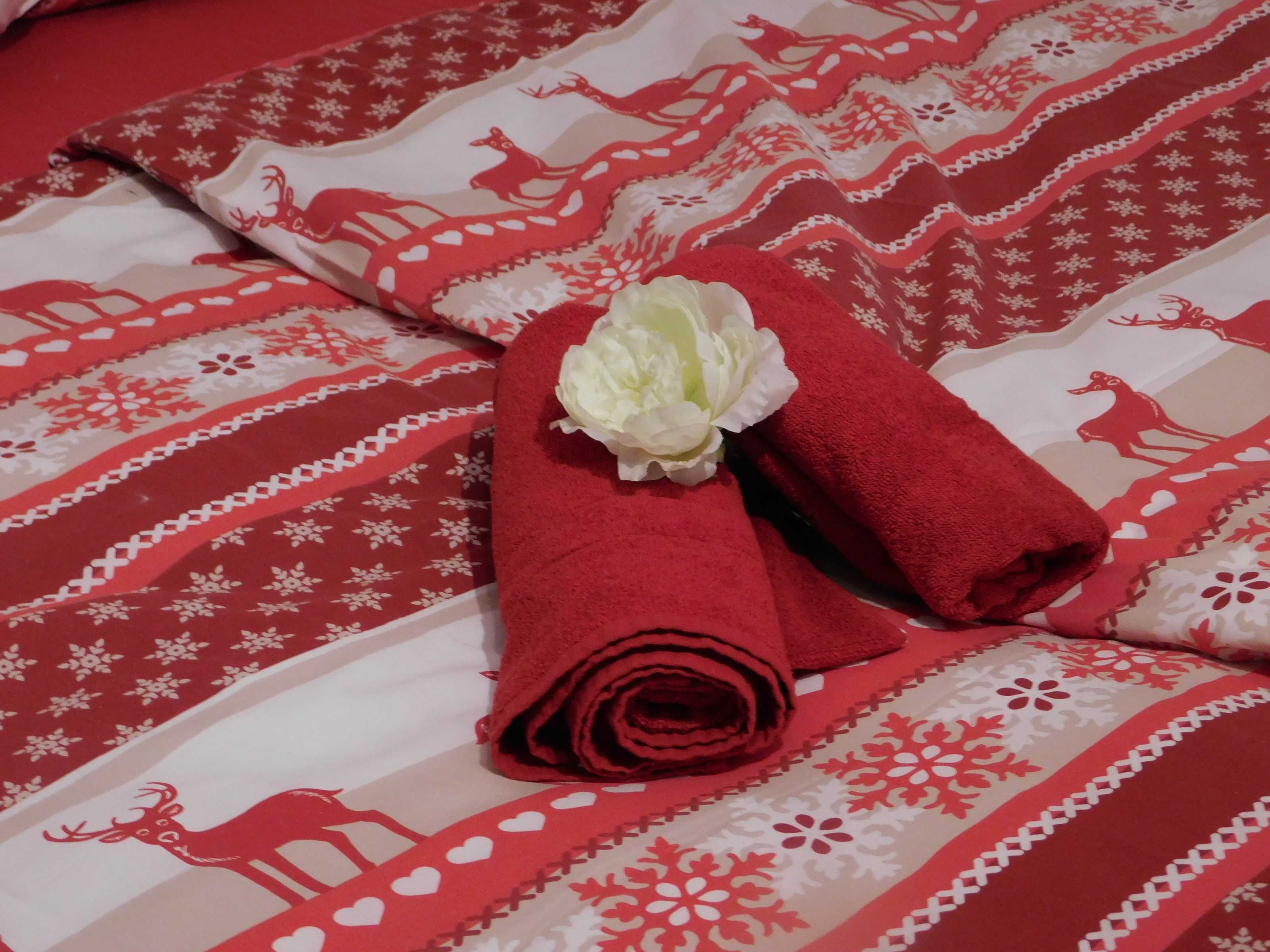 Ein Bett mit einer rot-weißen Bettdecke, ein gerolltes rotes Handtuch und eine weiße Blume.