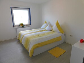 Een slaapkamer met een bed, een raam, een radiator en een kleine tafel met een rood voorwerp erop.