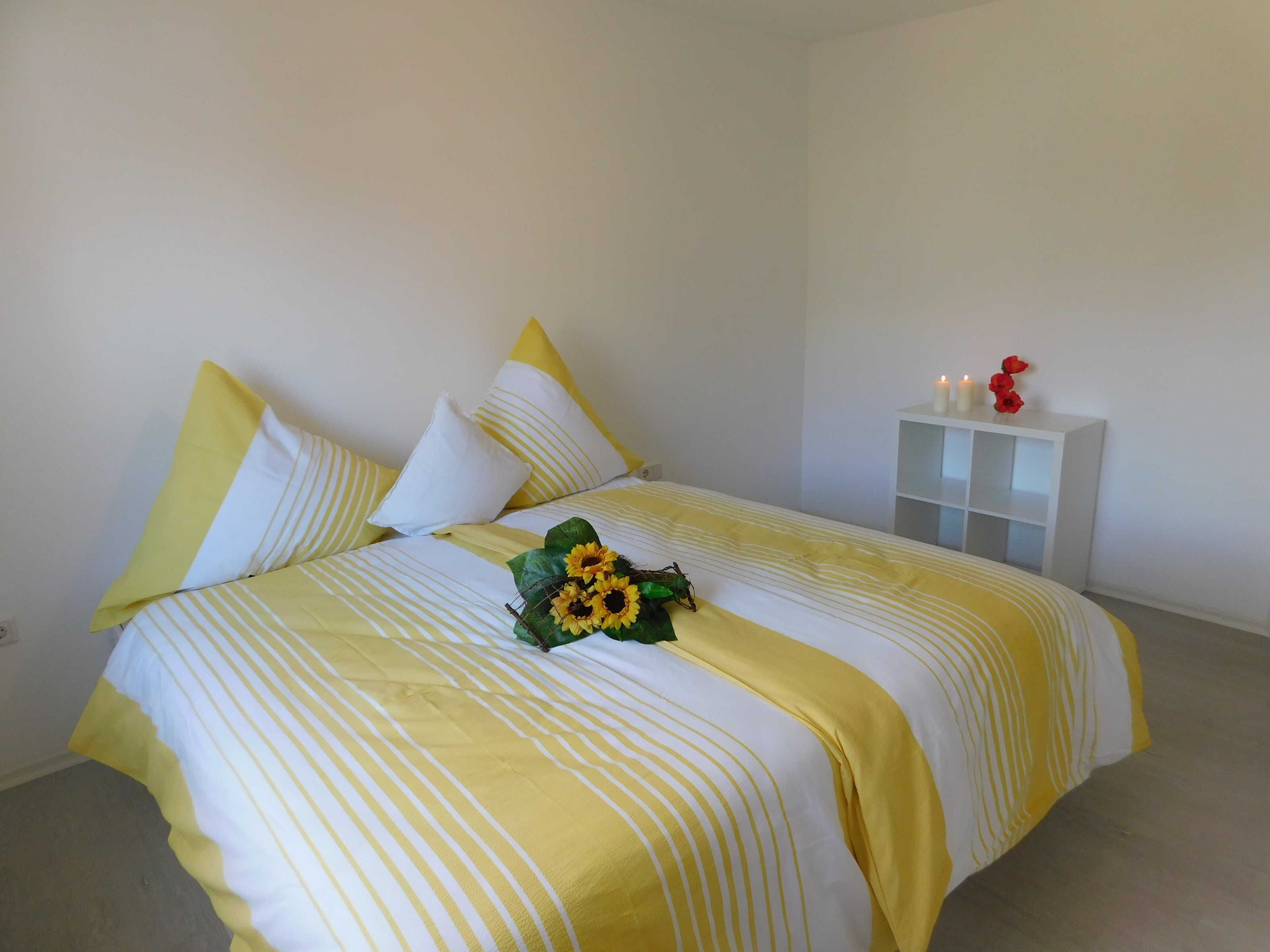 Ein Bett mit gelb-weiß gestreifter Bettwäsche, ein Sonnenblumenstrauß und ein weißer Nachttisch mit Kerzen.