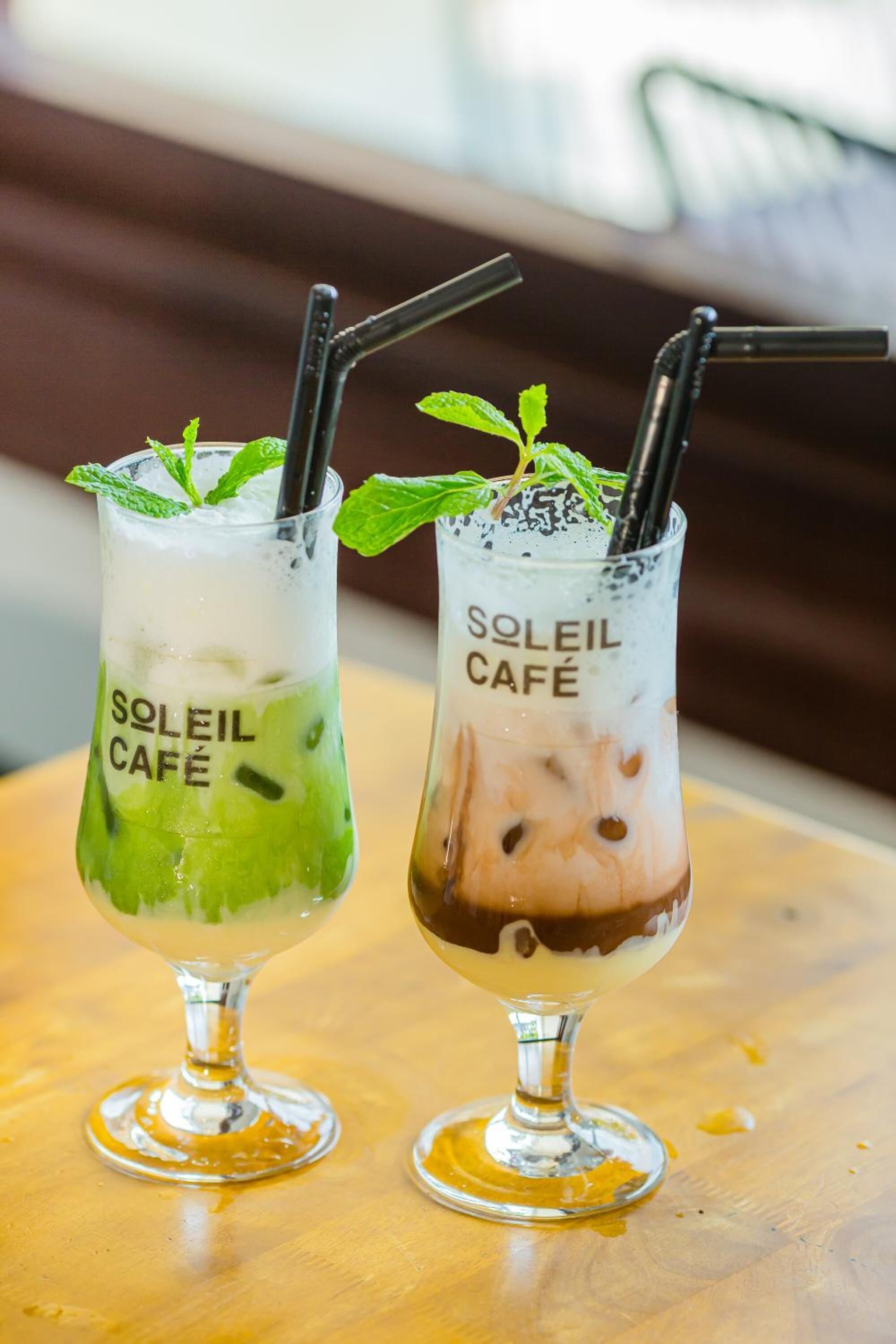 Two drinks in glasses with straws, one with a green beverage and mint, and the other with a brown beverage and mint, both labeled "SOLEIL CAFE".