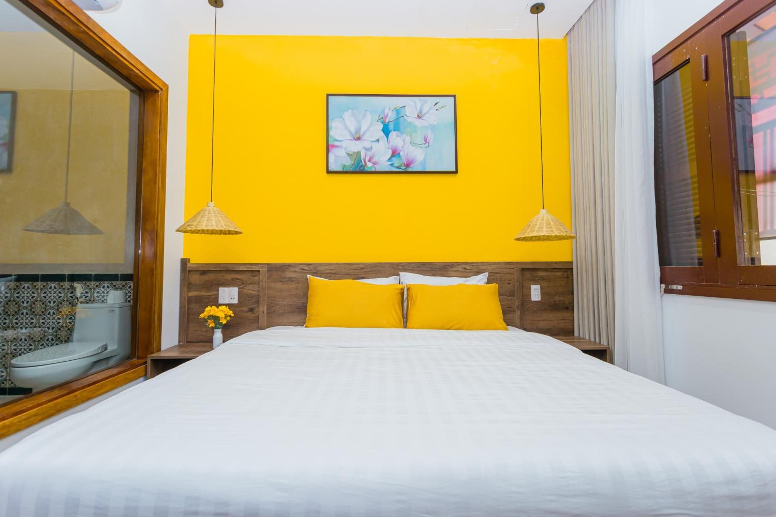 A hotel room with a bed, a painting on the wall, and a bathroom visible through a glass partition.
