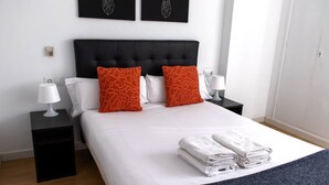 Ein ordentlich bezogenes Bett mit weißen Leinen und zwei orangefarbenen Kissen, ein schwarzer Nachttisch mit einer weißen Lampe und ein gefaltetes weißes Handtuch auf dem Bett.