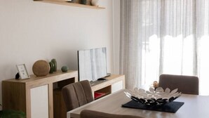 Ein modernes Wohnzimmer mit Flachbildfernseher, einem Esstisch mit Stühlen, einem Holzkonsole und dekorativen Accessoires sowie transparenten Vorhängen.
