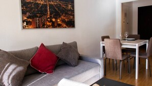 Ein Wohnzimmer mit grauem Sofa, roten und gemusterten Kissen, einem Esstisch mit Stühlen und einem Stadtbild an der Wand.