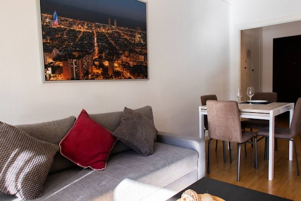 A living room with a grey sofa, red and patterned cushions, a dining table with chairs, and a cityscape painting on the wall.