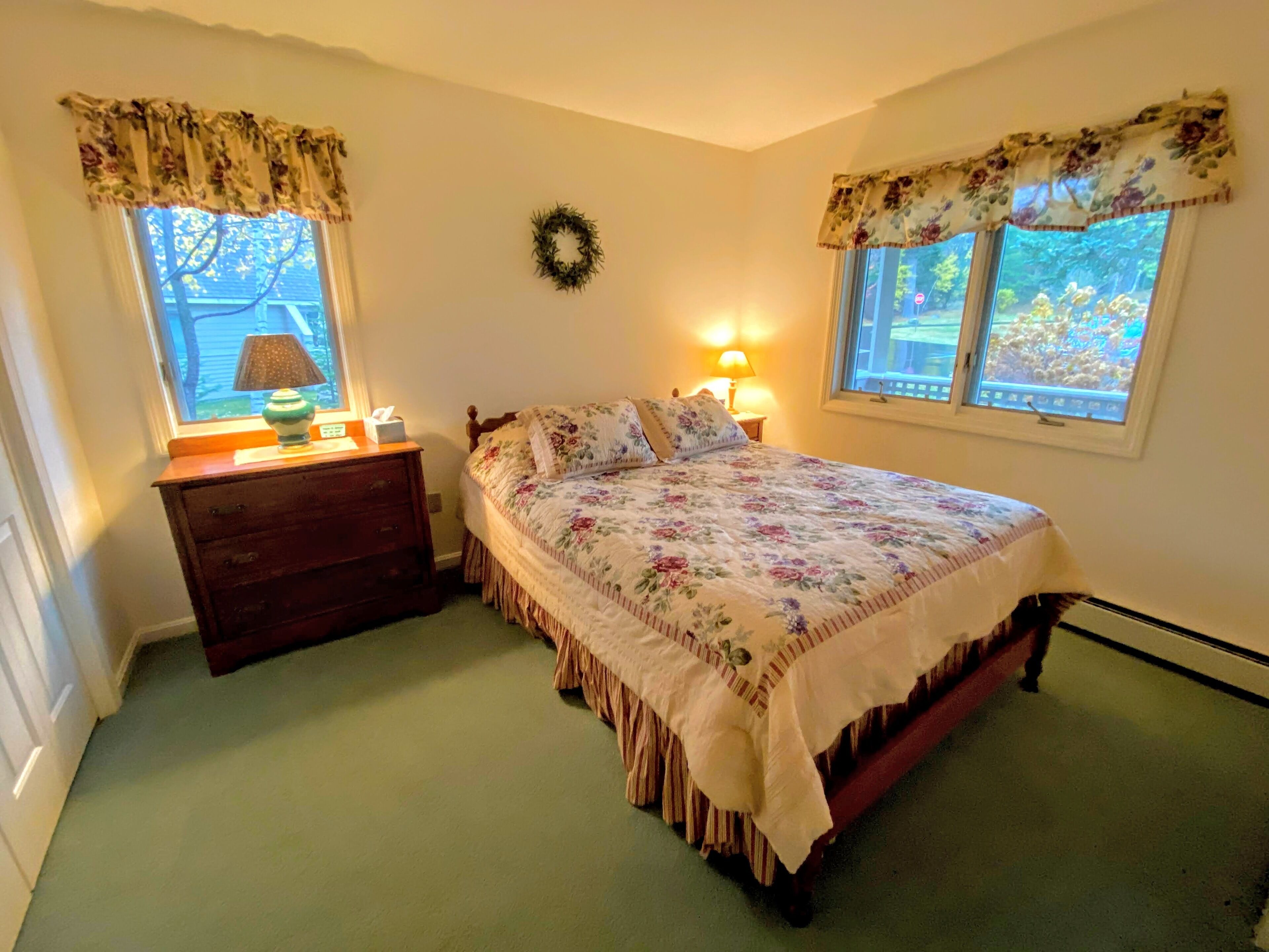 A bedroom with a bed, dresser, two windows with floral valances, a wall-mounted wreath, and a nightstand with a lamp.
