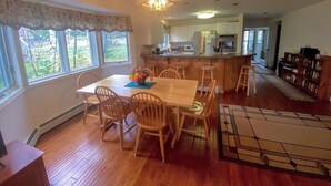 A spacious kitchen with a central island, wooden dining table with chairs, and a view of the outdoors through large windows.