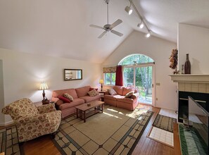 A cozy living room with a ceiling fan, a fireplace, and a large window overlooking a garden.