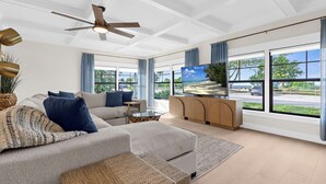 A spacious living room with a large sofa, a coffee table, and a TV on a cabinet. There are large windows with a view of the outdoors. - ⚓Bay Dreamin! Walk to the Beach, Pier, & Pine Ave! Heated Pool!⚓⛳🏖🌅 (Anna Maria)