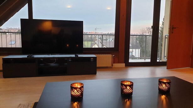 A modern living room with a large flat-screen TV on a black entertainment unit, two decorative candles on a table, and a view of a residential area through large windows.