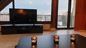 Ein modernes Wohnzimmer mit einem großen Flachbildfernseher auf einer schwarzen Unterhaltungseinheit, zwei dekorative Kerzen auf einem Tisch und Blick auf eine Wohngegend durch große Fenster.