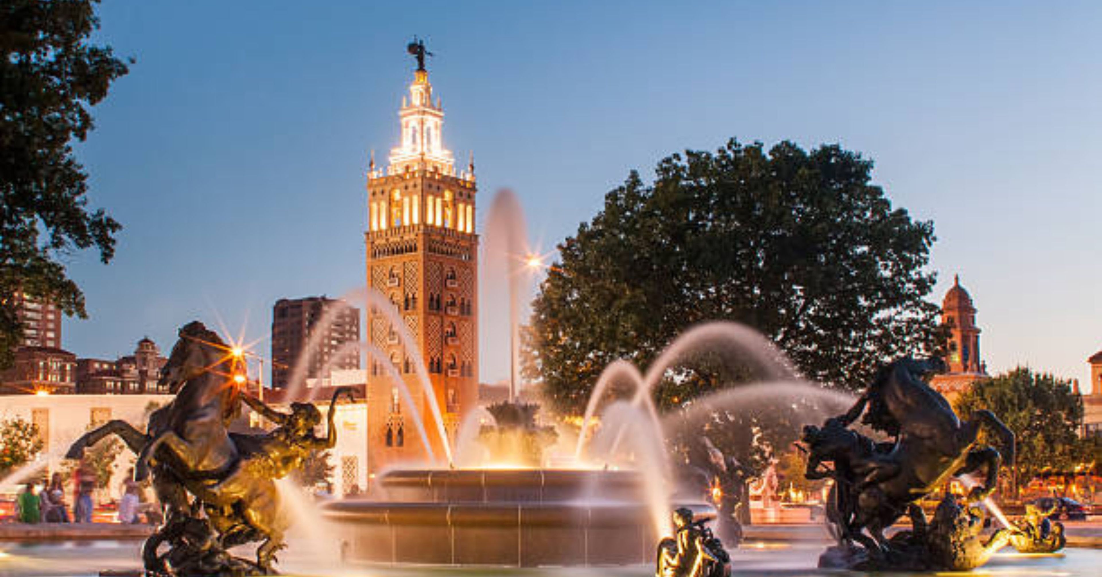 Une fontaine avec des jets d’eau, entourée de statues de chevaux et de cavaliers, avec un grand immeuble en arrière-plan.