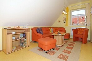 A compact living space with a sofa, ottoman, and a small round table. A bookshelf is against the wall, and a window with curtains is visible.