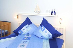 A bed with blue bedding and pillows, a small shelf with bottles and a model sailboat, and a crib in the corner.