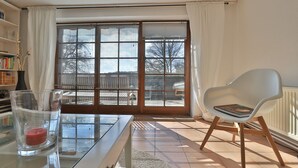 Un salon avec une table en verre, une chaise blanche, une bibliothèque et une grande porte-fenêtre coulissante donnant sur une terrasse.