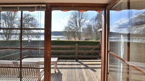 Vue depuis une pièce dotée de grandes fenêtres donnant sur une terrasse en bois et un lac.