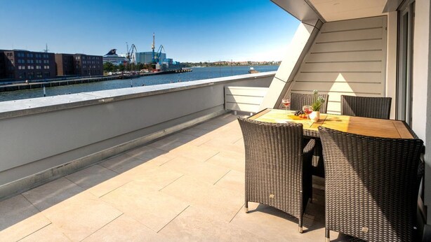 Un balcon avec une table et des chaises, donnant sur un front de mer où l’on aperçoit des bâtiments et des grues au loin.