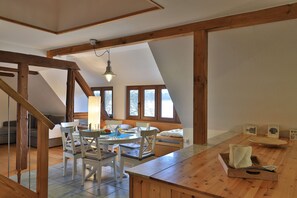 A spacious loft-style room with a high ceiling, exposed wooden beams, a dining area with a table and chairs, and large windows offering views of the outdoors.