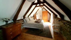 A bedroom with a sloped ceiling, wooden beams, a bed with patterned bedding, a dresser, a vase with flowers, and a stone wall. - In the grounds of a manor house (Saint Hélen)