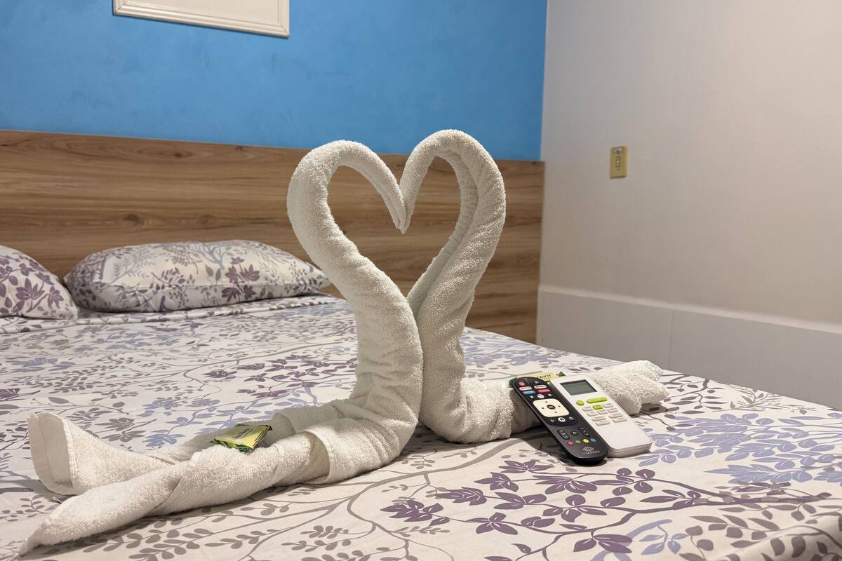 Un lit recouvert d’une couvre-lit à motifs floraux, une sculpture en forme de cœur faite avec une serviette et une télécommande posées sur le lit.