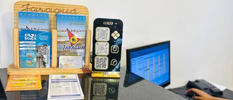 A reception desk with a wooden display board, two brochures, a QR code device, and a computer monitor.