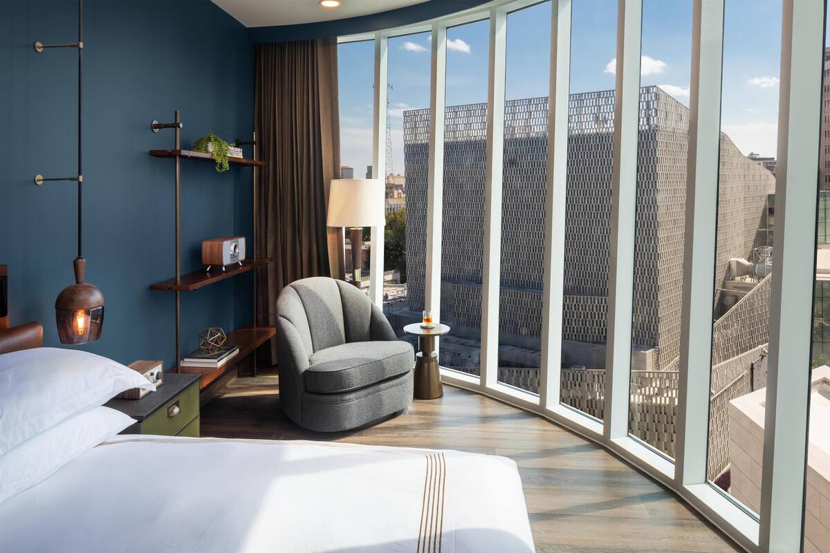 Une chambre d’hôtel moderne dotée d’un grand lit, d’un coin salon avec un fauteuil et une table d’appoint, d’une lampe, et offrant une vue sur la ville grâce à de grandes fenêtres.
