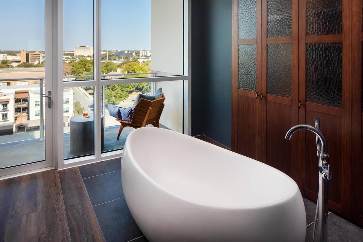 Une salle de bain moderne avec une baignoire sur pieds, une chaise en bois avec un coussin et une grande fenêtre donnant sur la ville.