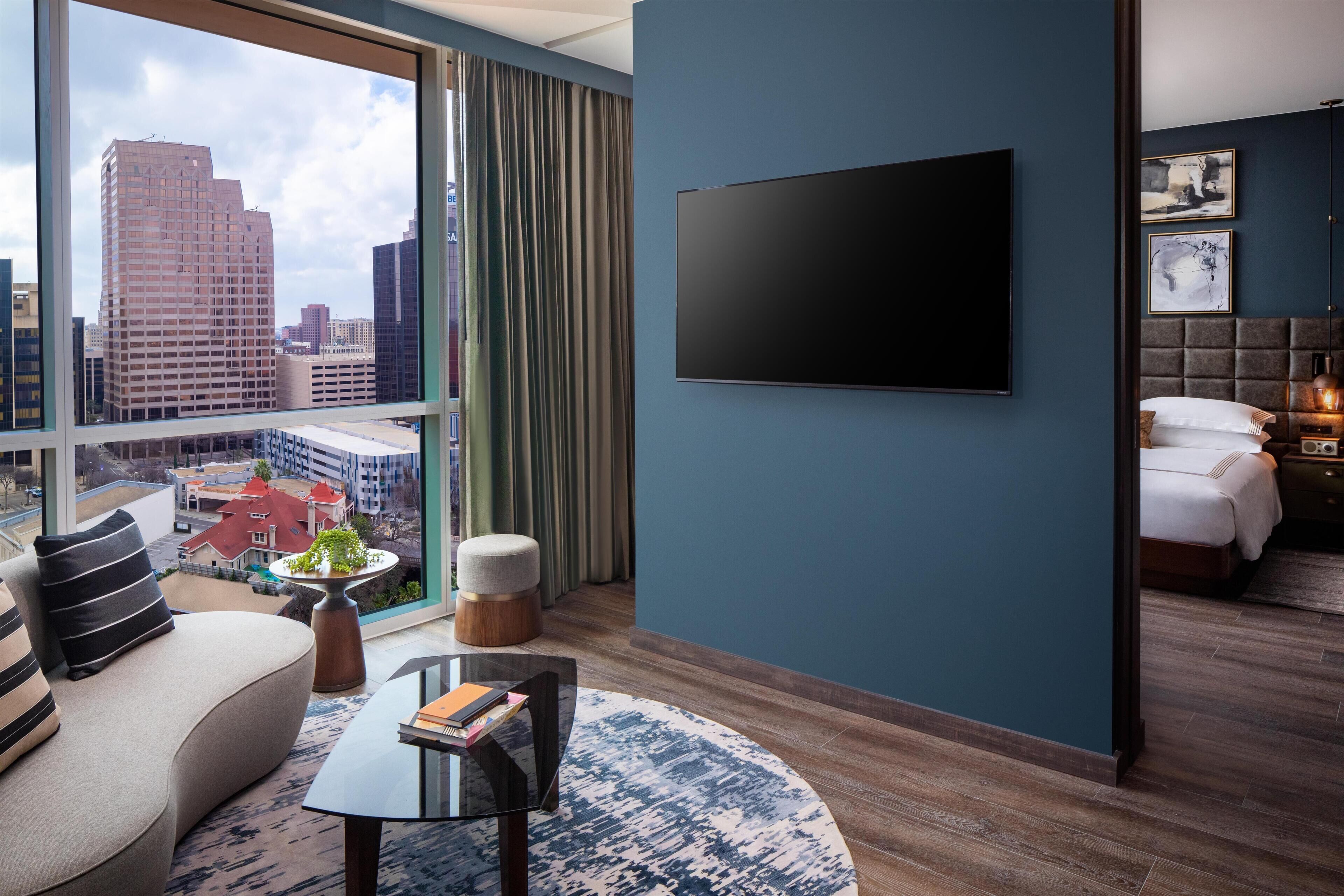 A modern hotel room with a large window, a flat-screen TV, a sofa, a coffee table, and a bed with a headboard.