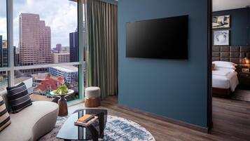 A modern hotel room with a large window, a flat-screen TV, a sofa, a coffee table, and a bed with a headboard.