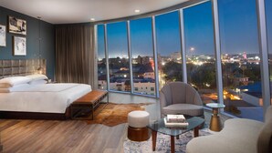 Una habitación de hotel moderna con una cama grande, una zona de estar con una silla y una mesa, y un ventanal con vistas a la ciudad.