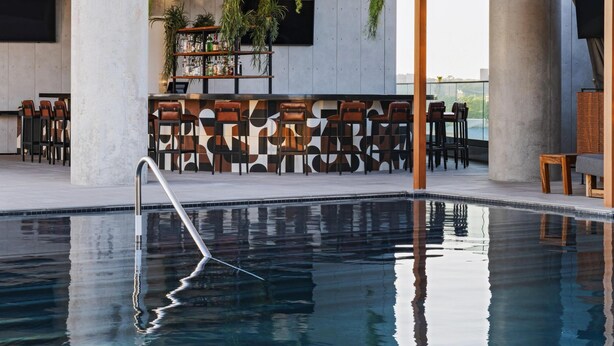 Zona moderna de piscina interior con agua cristalina, rodeada de asientos y una zona de bar con plantas y un televisor.