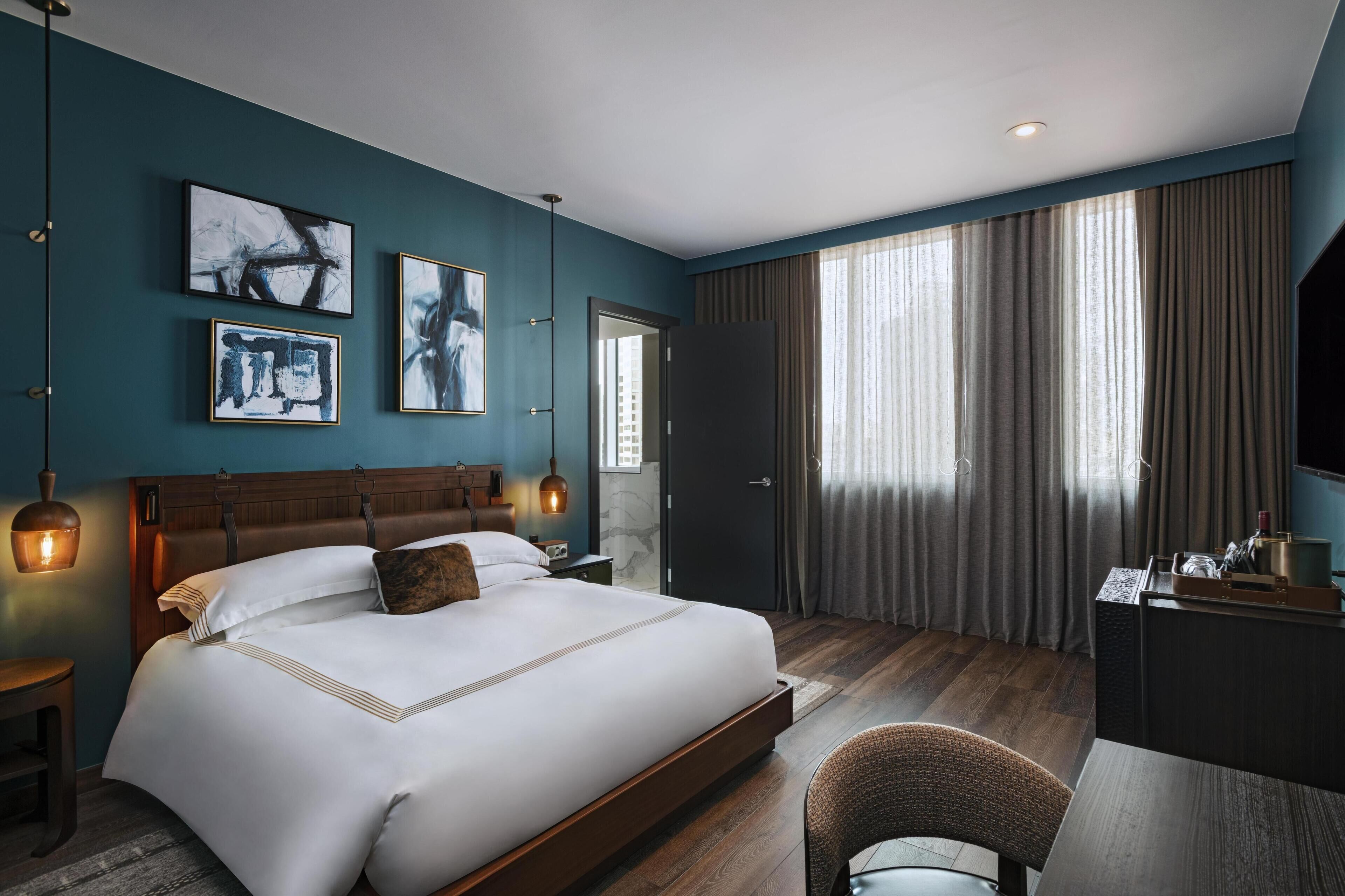 Habitación de hotel moderna con una cama grande, un escritorio y una silla. Hay obras de arte en las paredes y una ventana con cortinas.